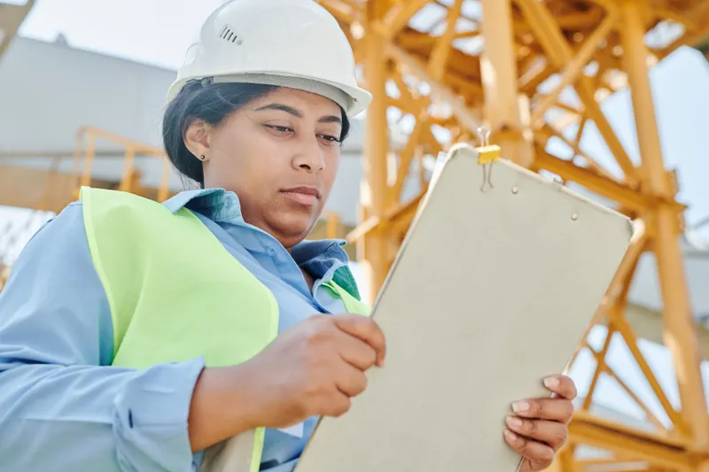 Confident female construction worker inspecting site with clipboard and helmet