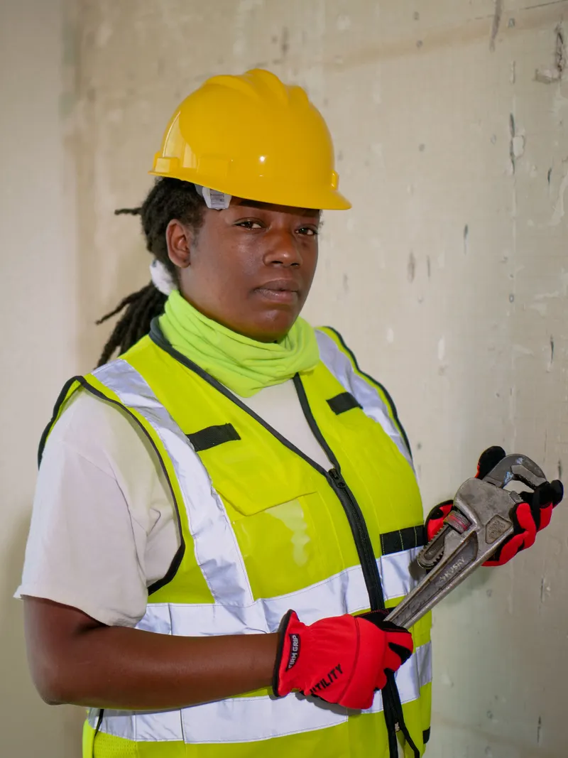 Confident handywoman wearing safety gear and holding a plumber's wrench indoors.